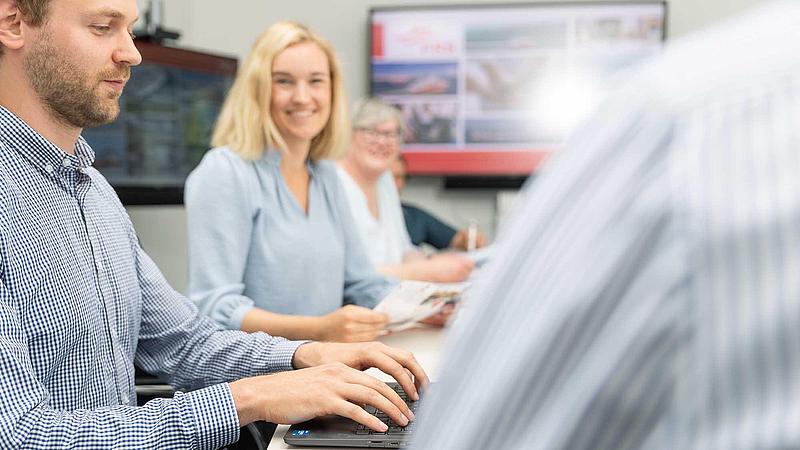 A man typing ona notebook and two women in a meeting situation with a screen in the background